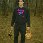 Person wearing Bunny Man Sweatshirt with neon pink text and graphic of a rabbit figure holding a small dog and an axe outdoors