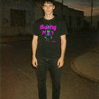 Man wearing black Bunny Man t-shirt with neon purple text and surreal bunny character standing on a dimly lit street at dusk