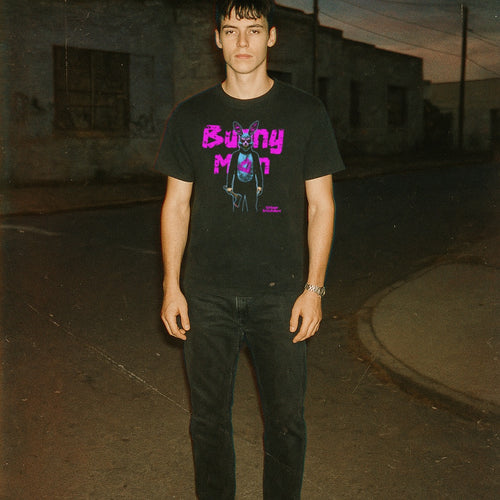 Man wearing black Bunny Man t-shirt with neon purple text and surreal bunny character standing on a dimly lit street at dusk