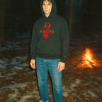 Person wearing a dark hoodie with red graphics and blue jeans standing near a campfire at night.