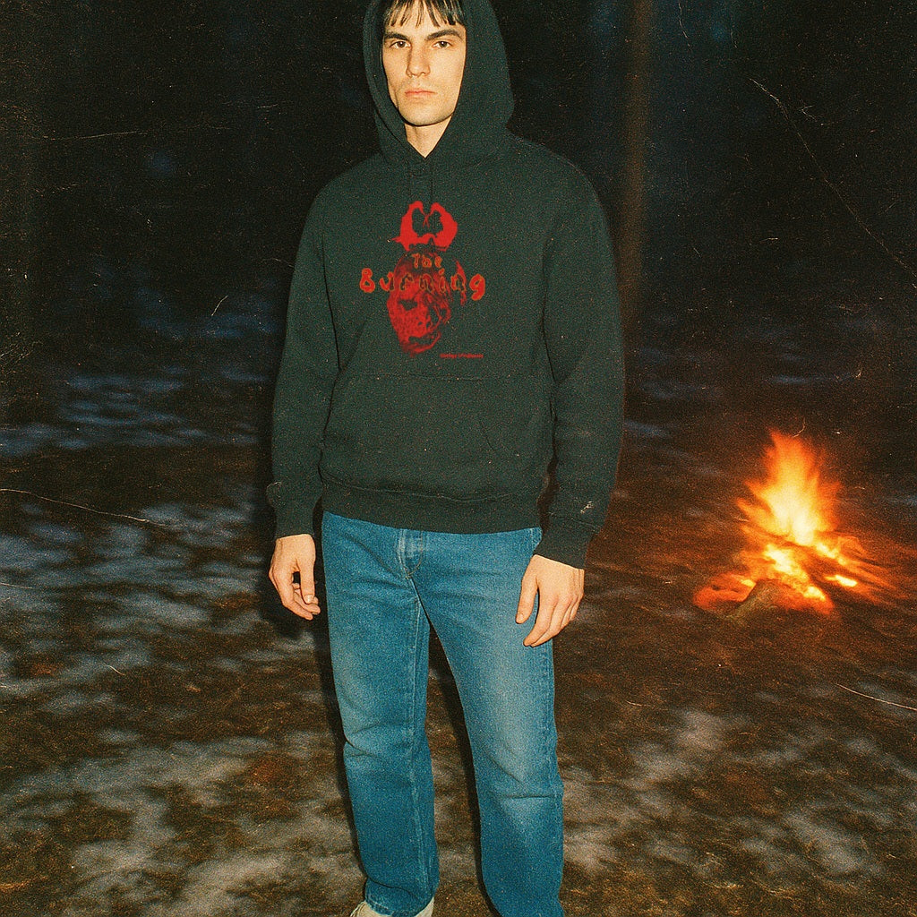 Person wearing a dark hoodie with red graphics and blue jeans standing near a campfire at night.