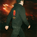 Person walking wearing Cannibal Holocaust hoodie (rear print) with bold red graphic and yellow text on black fabric at night