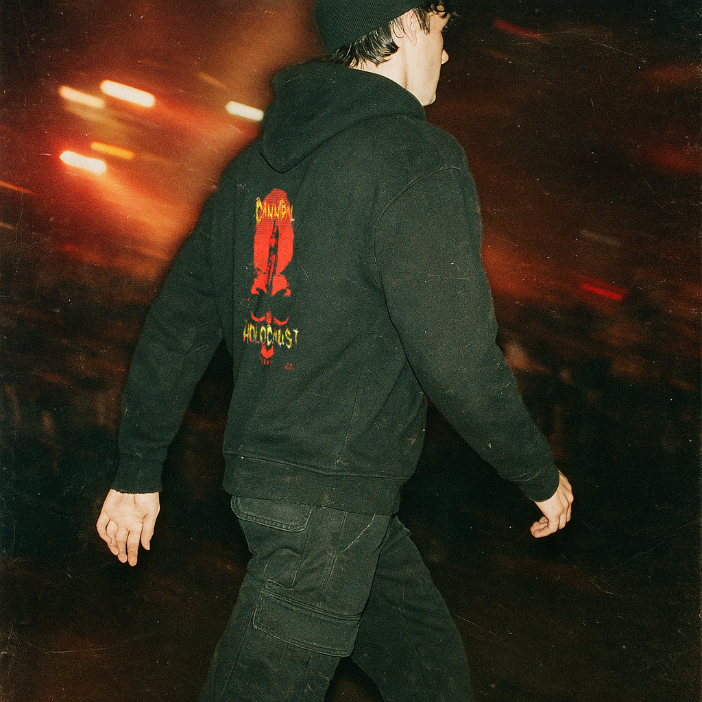 Person walking wearing Cannibal Holocaust hoodie (rear print) with bold red graphic and yellow text on black fabric at night