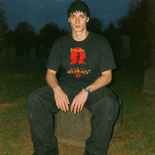 Model wearing black Cannibal Holocaust t-shirt with striking red and yellow graphic sitting outdoors at dusk