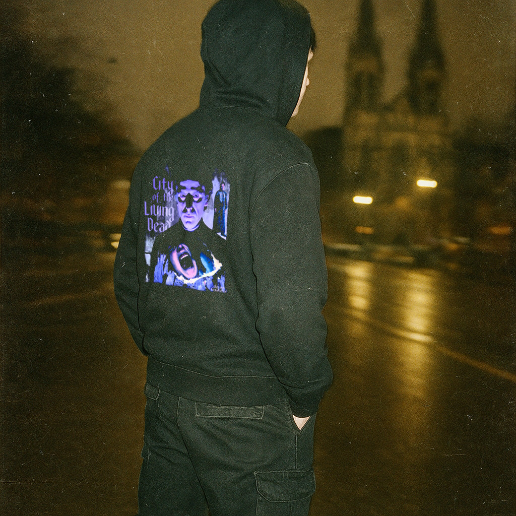 Person wearing black hoodie with purple graphic and text City of the Living Dead rear print standing on wet street at night