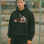 Person wearing a black hoodie with graphic design in front of a chain-link fence.