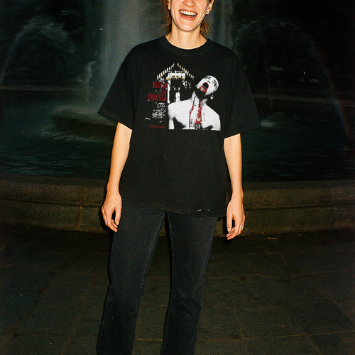 Person wearing a black t-shirt with graphic design, standing in front of a fountain at night.
