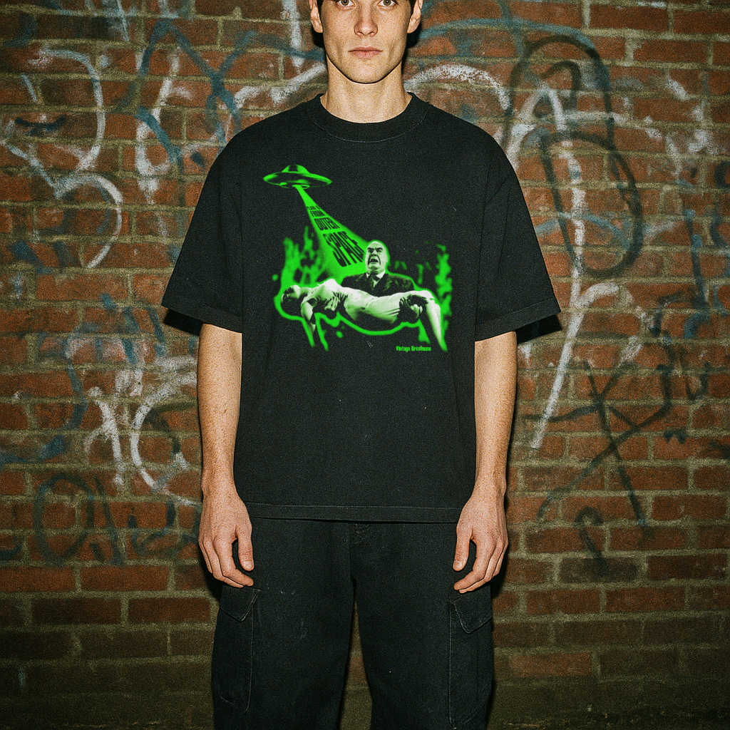 Person wearing a black t-shirt with a green graphic design in front of a graffiti-covered brick wall.