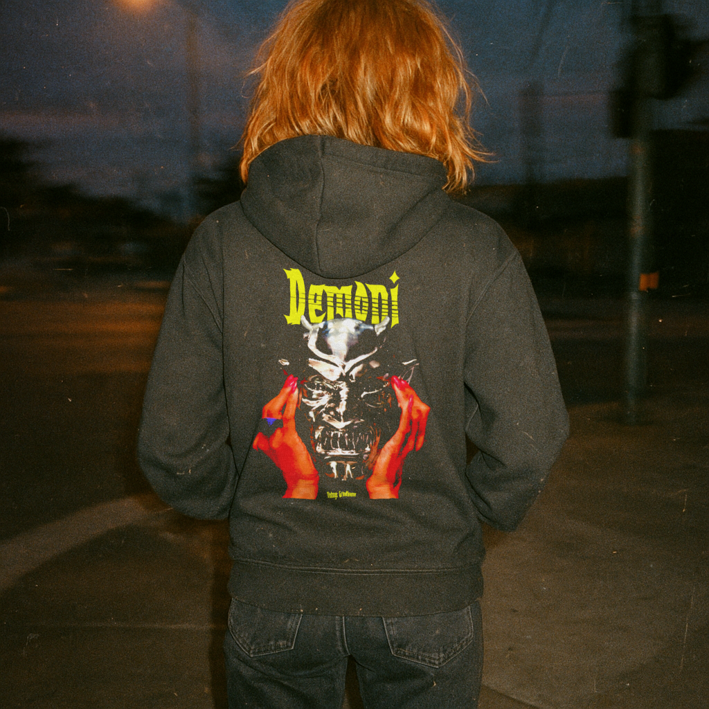 Person outdoors wearing Demoni hoodie (rear print) with vivid horror mask graphic and yellow lettering on black fabric at night