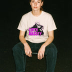 Person seated wearing cream Edith Massey - Legendary Lady of Grindhouse Cinema t-shirt with bold pink and black design at night