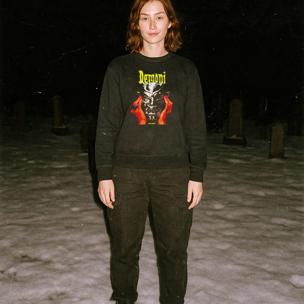 Person standing outdoors at night wearing Demoni aka Demons sweatshirt with bold yellow text and fiery red hands graphic