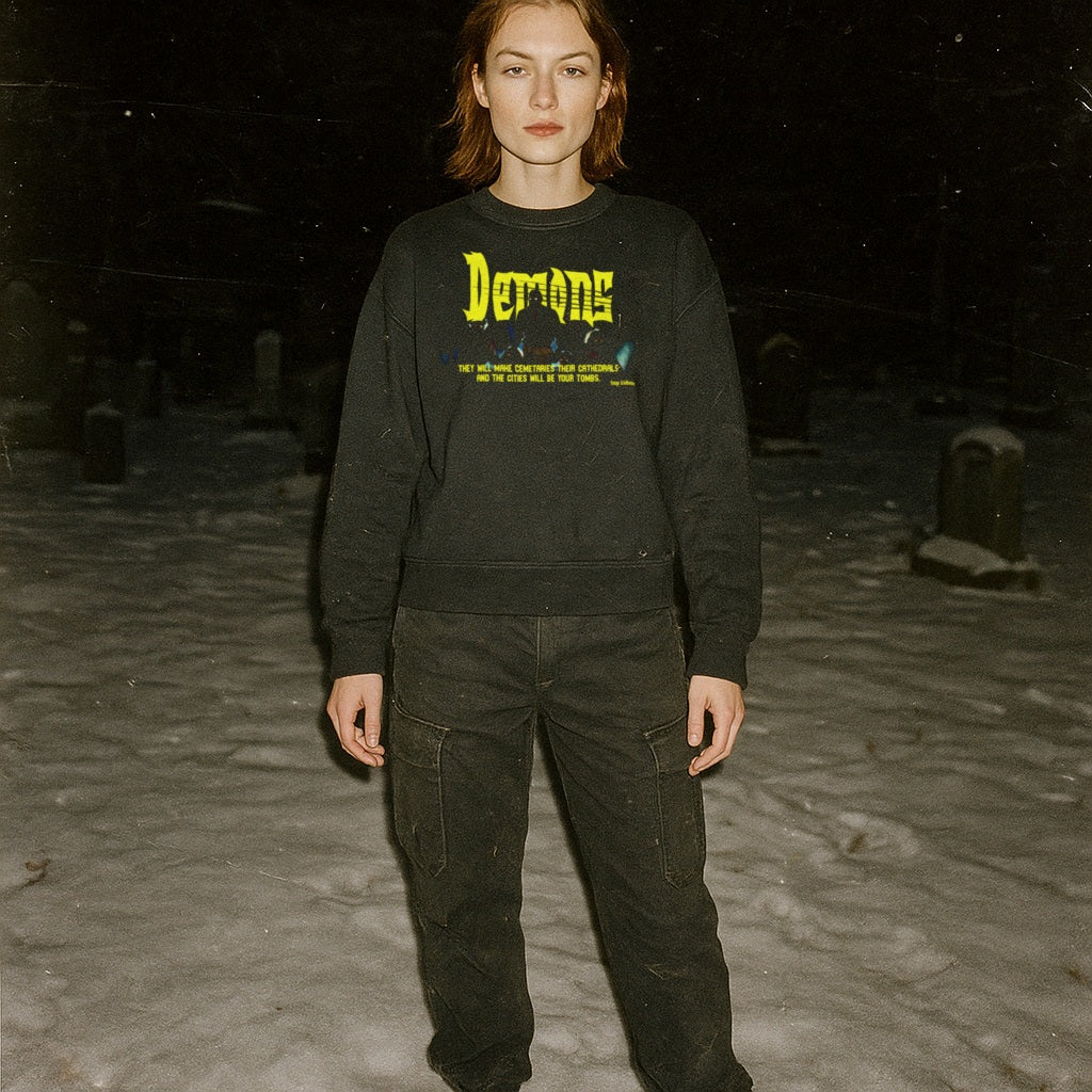 Person standing in snowy graveyard wearing Demons aka Demoni sweatshirt with bold yellow text and dark graphic on black fabric