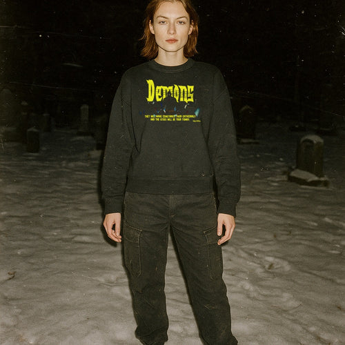 Person standing in snowy graveyard wearing Demons aka Demoni sweatshirt with bold yellow text and dark graphic on black fabric