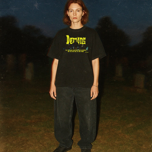 Person wearing a black t-shirt with 'Demons' text in a dark, possibly cemetery setting.