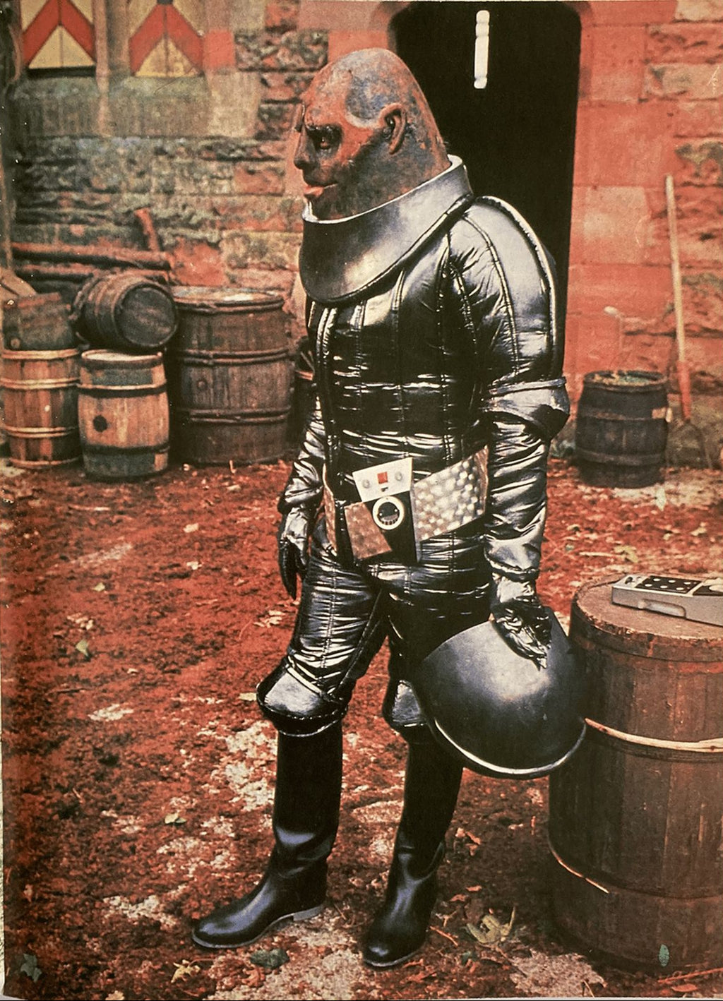 Person in full body armor standing in a rustic setting with barrels and stone walls.