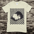 Beige t-shirt with a graphic design of a face on a checkered background against a brick wall.