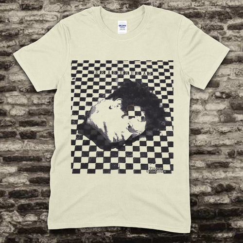 Beige t-shirt with a graphic design of a face on a checkered background against a brick wall.