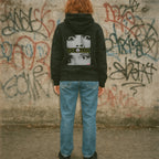 Person wearing a black hoodie with a graphic design in front of a graffiti-covered wall.