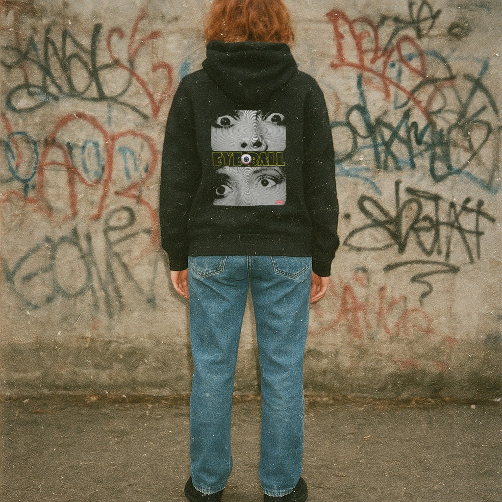 Person wearing a black hoodie with a graphic design in front of a graffiti-covered wall.