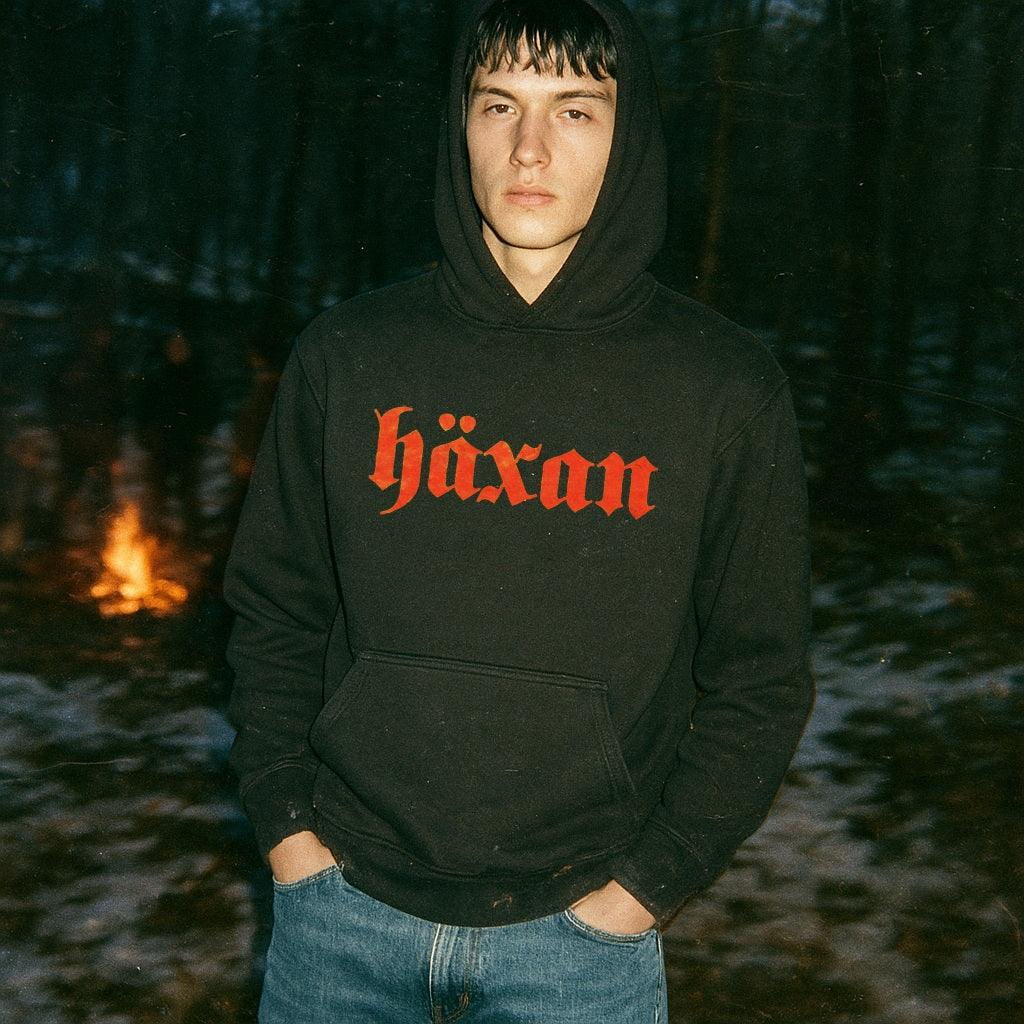 Person wearing a black hoodie with 'häxan' in red text, standing in a forest with a fire in the background.