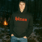 Person wearing a black hoodie with 'häxan' in red text, standing in a forest with a fire in the background.