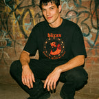 Man wearing a black t-shirt with red text and graphics against a graffiti-covered wall.