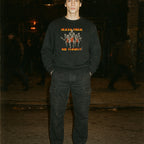 Person wearing a black sweatshirt with graphic design and text, standing in a dark, possibly urban setting.