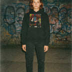 Person wearing a black hoodie with a graphic design in front of a graffiti-covered wall.