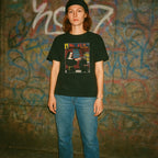 Person wearing a black t-shirt with a graphic design in front of a graffiti-covered wall.