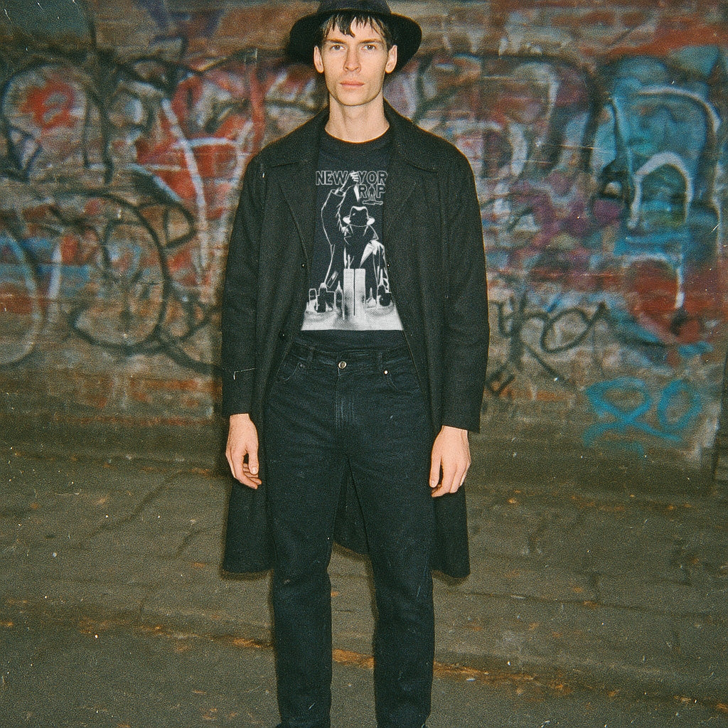 Model wearing black coat and hat with The New York Ripper graphic t-shirt against graffiti-covered wall background