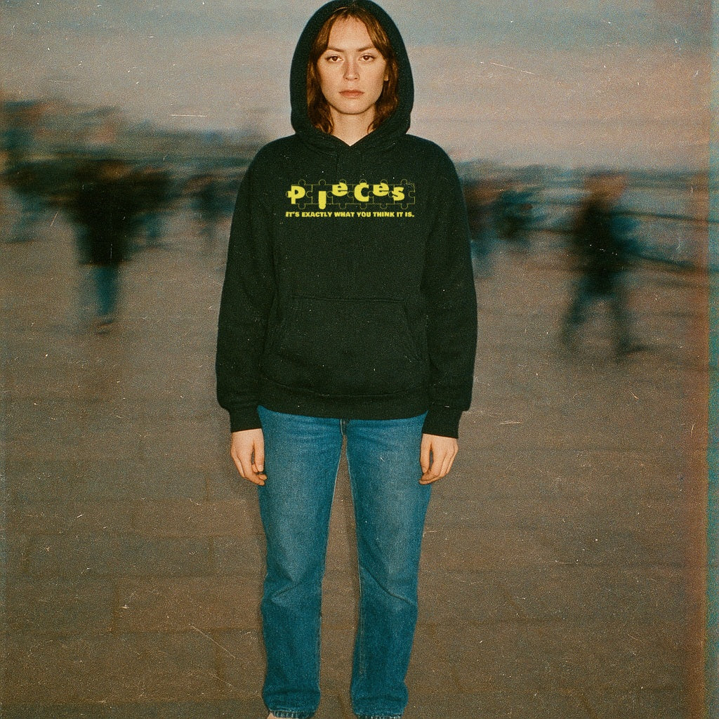 Person wearing Pieces - Crime Scene Edition hoodie with yellow text on black fabric standing outdoors with blurred background figures