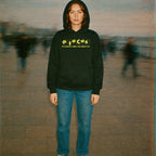 Person wearing Pieces - Crime Scene Edition hoodie with yellow text on black fabric standing outdoors with blurred background figures