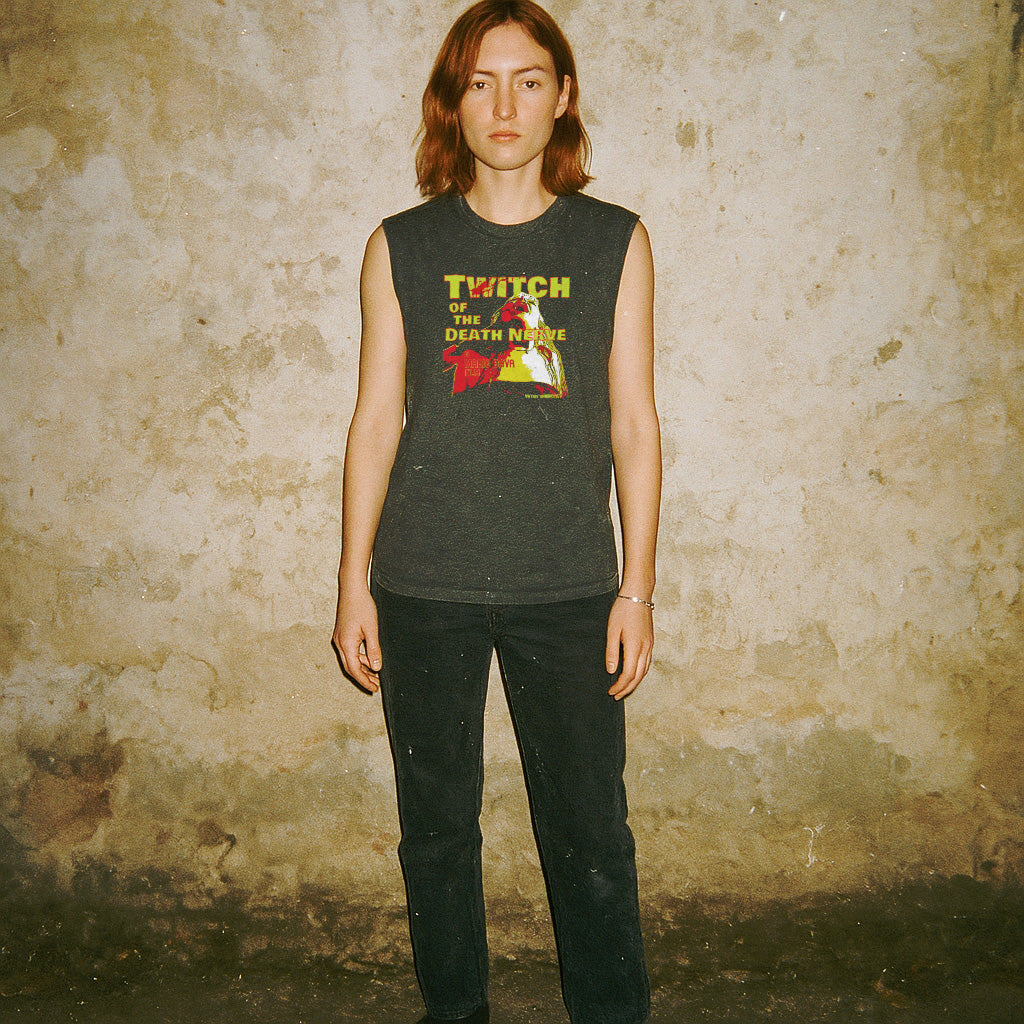 Woman modeling Twitch of the Death Nerve muscle tank with vintage horror-inspired graphic on dark fabric against textured wall