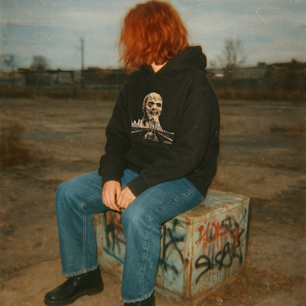 Person wearing Zombi 2 hoodie with skull and cityscape design sitting on graffiti-covered concrete block outdoors