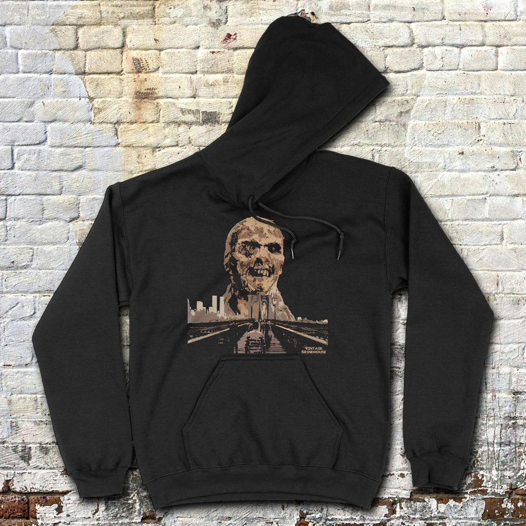 Zombi 2 hoodie with eerie zombie face and city skyline graphic on black fabric laid flat against brick wall background