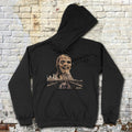 Zombi 2 hoodie with eerie zombie face and city skyline graphic on black fabric laid flat against brick wall background