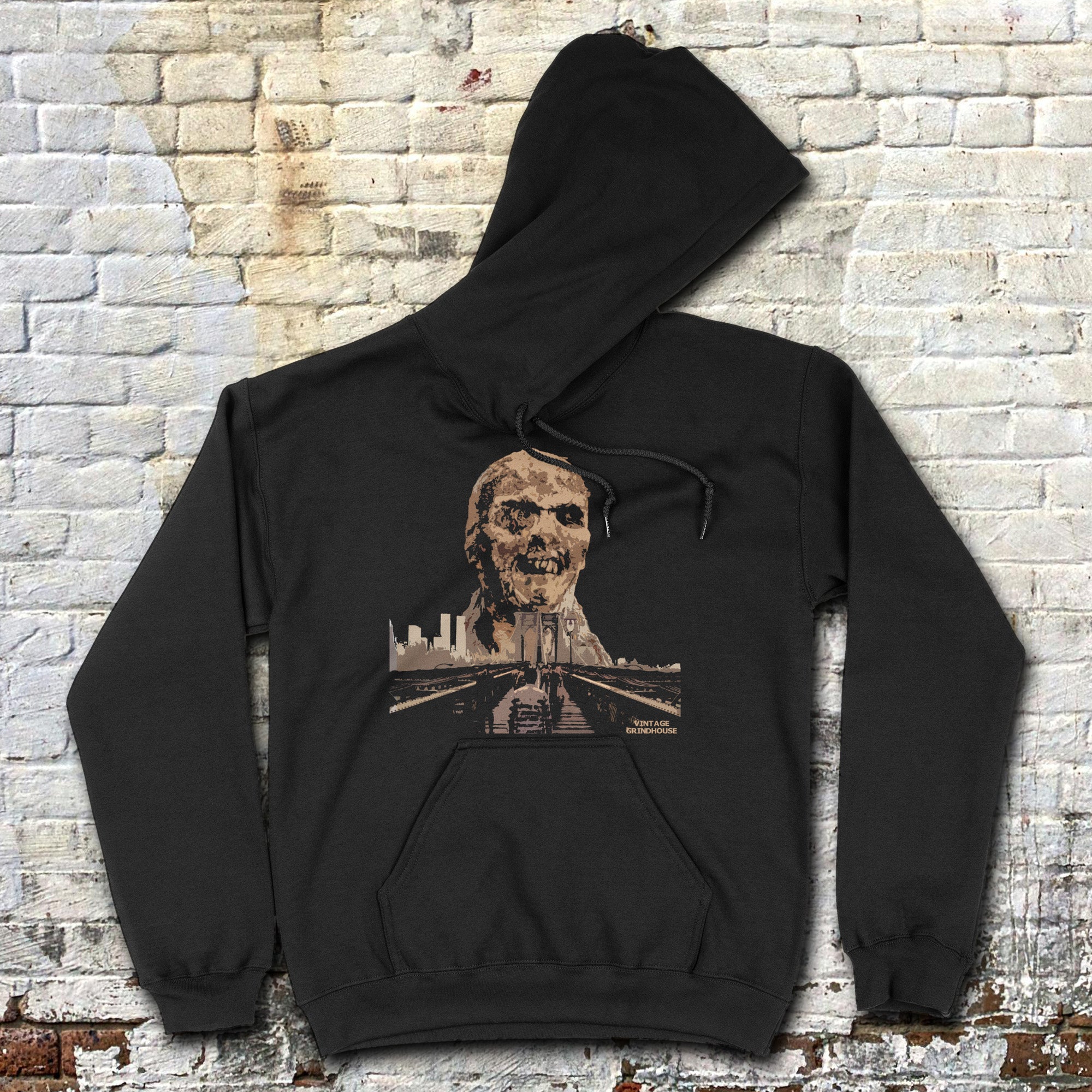 Zombi 2 hoodie with eerie zombie face and city skyline graphic on black fabric laid flat against brick wall background